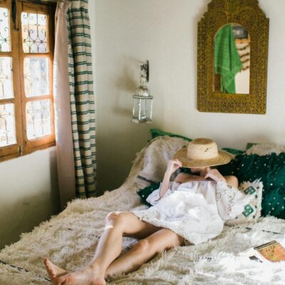 Anonymous woman in white dress covering face with straw hat while lying on bed in exotic hotel room