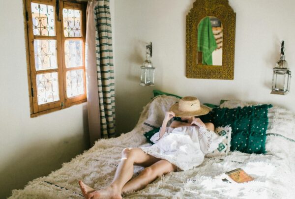 Anonymous woman in white dress covering face with straw hat while lying on bed in exotic hotel room