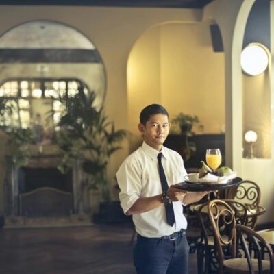 Confident young ethnic waiter in elegant clothes holding tray with food and drinks and looking at camera while serving tables in stylish restaurant