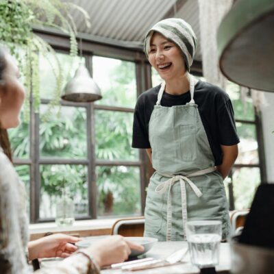 Happy laughing young Asian waitress in uniform standing near table and talking with female client while working in spacious light contemporary restaurant against big window and lush park