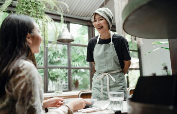 Happy laughing young Asian waitress in uniform standing near table and talking with female client while working in spacious light contemporary restaurant against big window and lush park